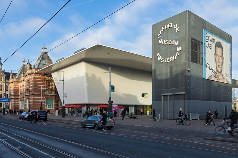 Street view of Stedelijk Museum Amsterdam showing the contrast between the historic brick building and the modern white bathtub wing extension.