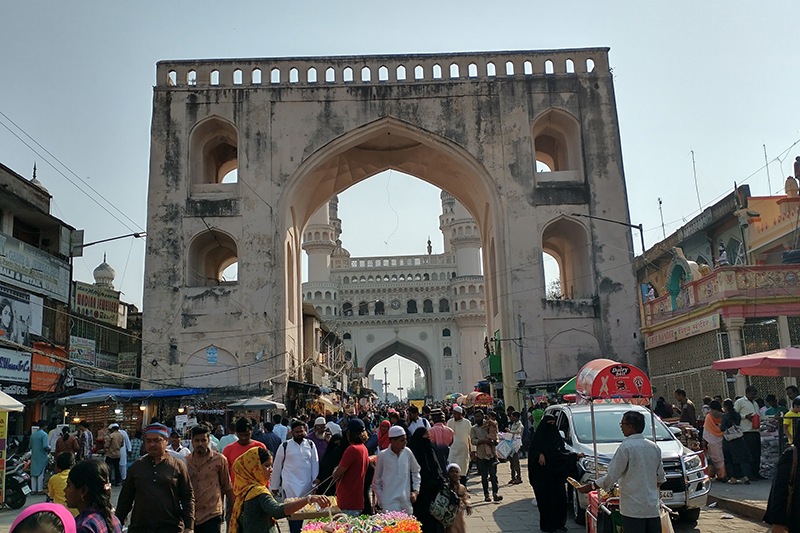 A busy street lined with market stalls and traditional shops, framed by the large historic stone archway of Machli Kaman under a clear sky.